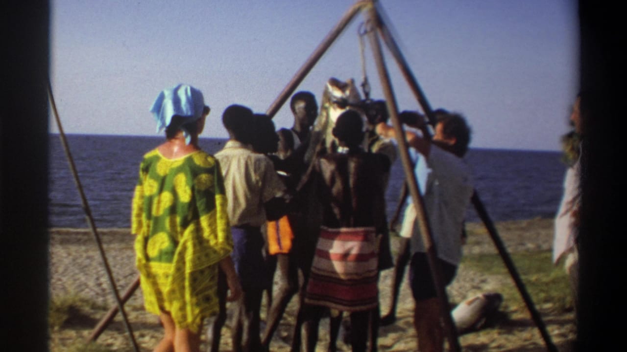 Adults in patterned and light clothing stand near a suspended large fish on a Nigerian shoreline in this 1969 home-movie frame.