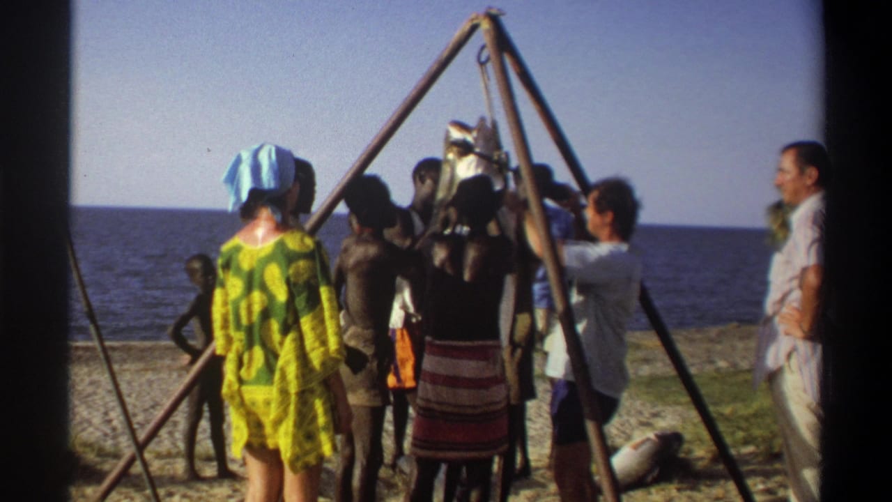1969 Nigeria 8mm frame with a woman in a yellow green patterned dress near a beach scaffold suspending a large fish.