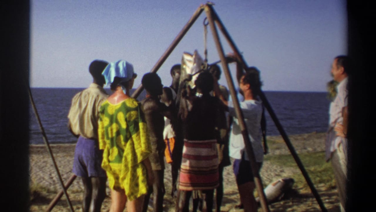A 1969 Nigeria home-movie frame shows adults gathered around a large fish suspended from a wooden A-frame beside the sea.