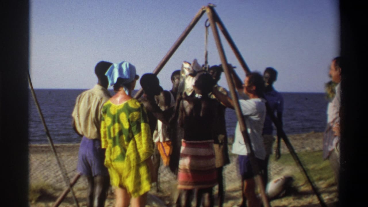 1969 Nigeria 8mm frame showing adults on a beach around a large fish suspended from a tripod, with ocean behind them.