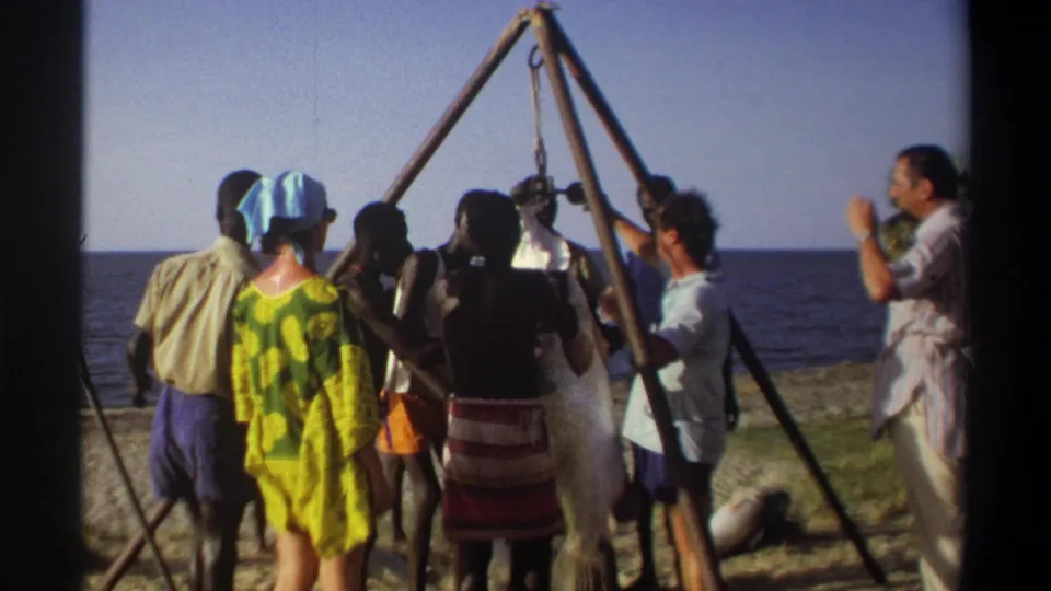 Adults in beach clothing and patterned dress stand around a suspended fish on a Nigerian shoreline in 1969 home-movie footage.