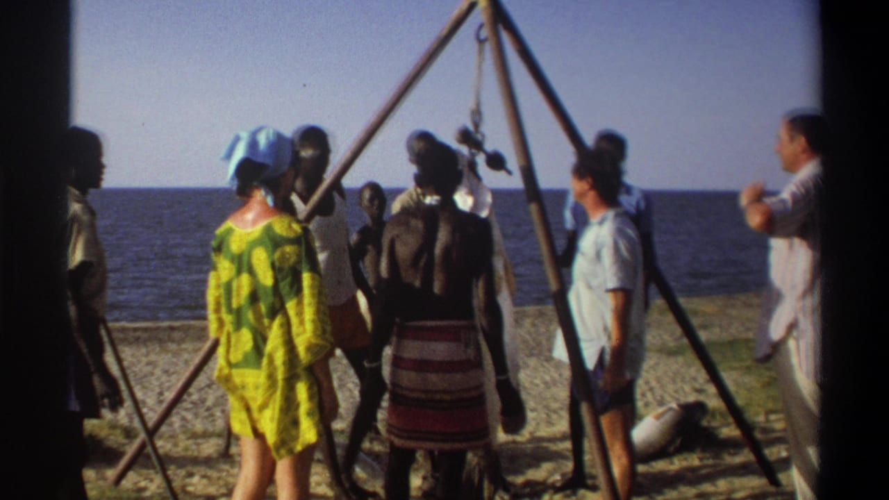 Rear view of adults on a Nigerian beach in 1969, gathered near a tripod with a large hanging fish against the sea.