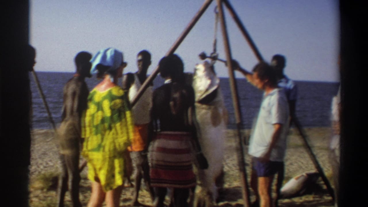 Final 1969 Nigeria beach frame with adults around a wooden tripod and large suspended fish, filmed in soft 8mm color.