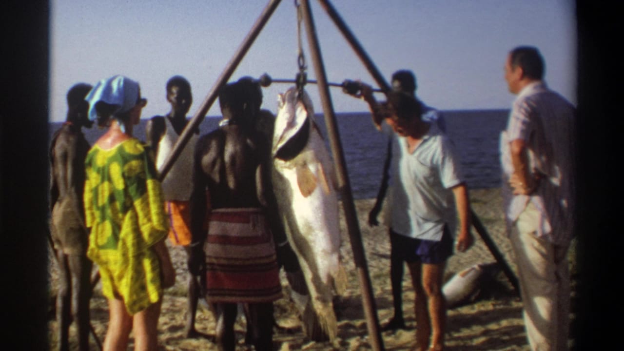 Nigeria 1969 8mm frame showing adults on a sandy beach beside a large fish suspended from a wooden tripod near the ocean.