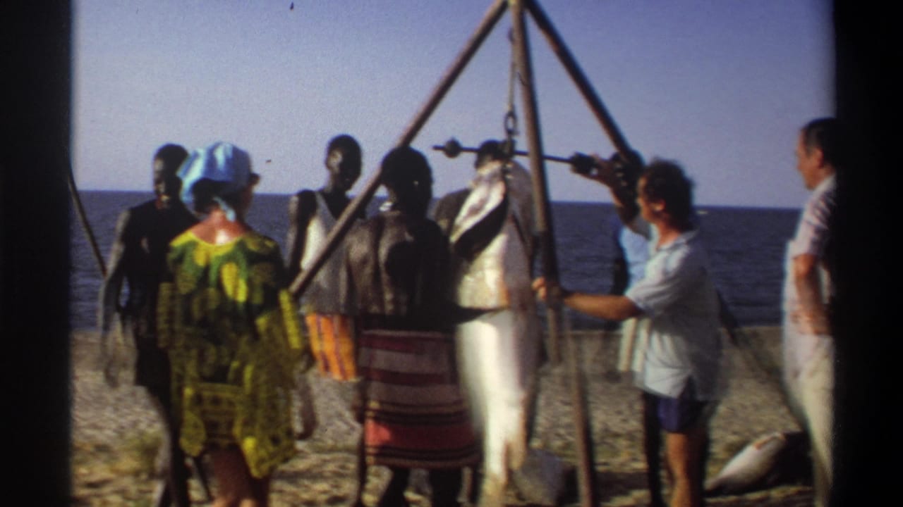 Nigeria 1969 beach scene with several adults gathered around a large hanging fish as a man in a light shirt adjusts the setup.