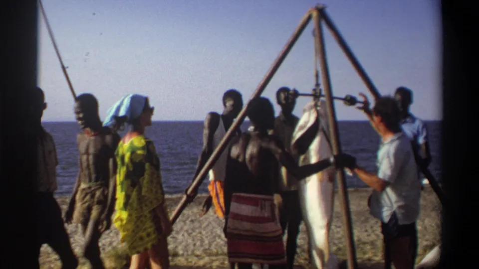 Nigeria 1969 8mm frame with adults beside a suspended large fish, the sea behind them and strong sun across the beach.