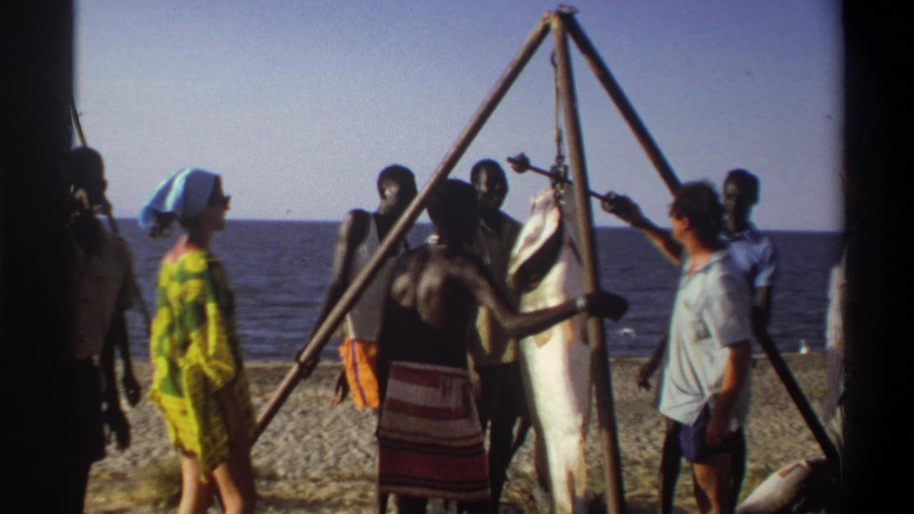 Nigeria 1969 frame showing a beach gathering around a large fish suspended from a tripod, with adults visible against the ocean horizon.
