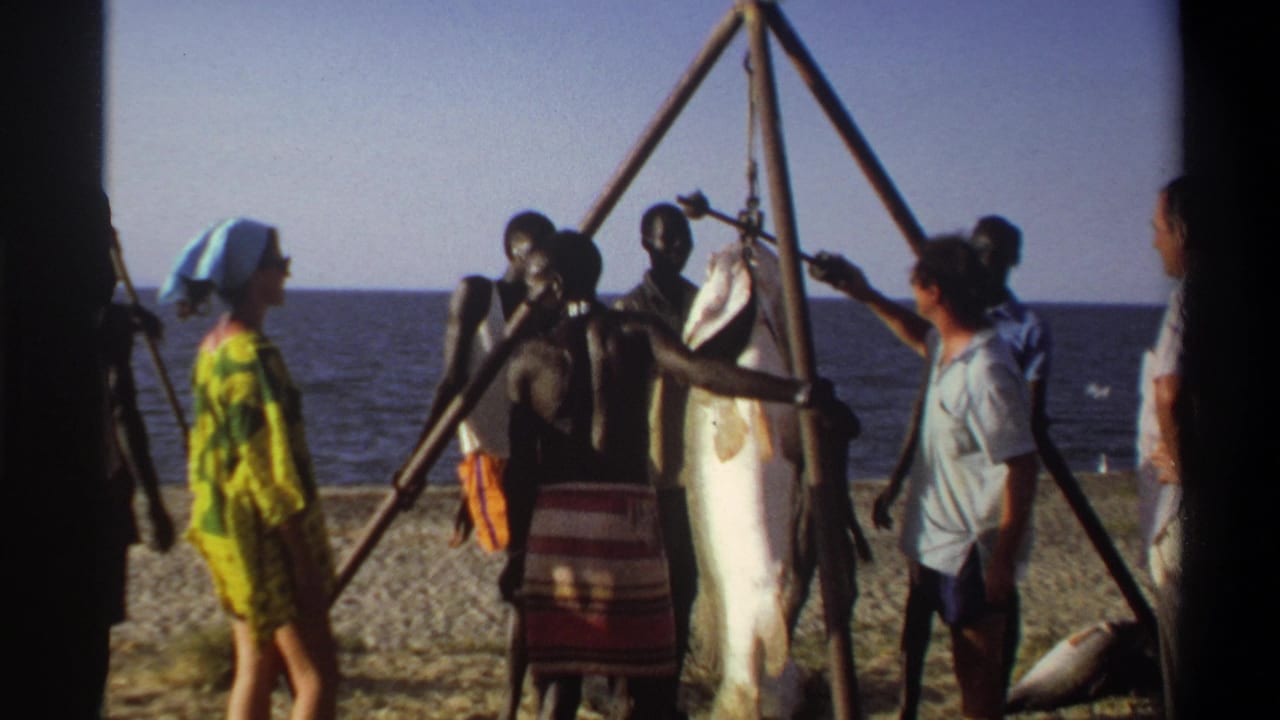 1969 Nigeria beach scene with people gathered around a large fish suspended from a triangular wooden pole rig near the sea.