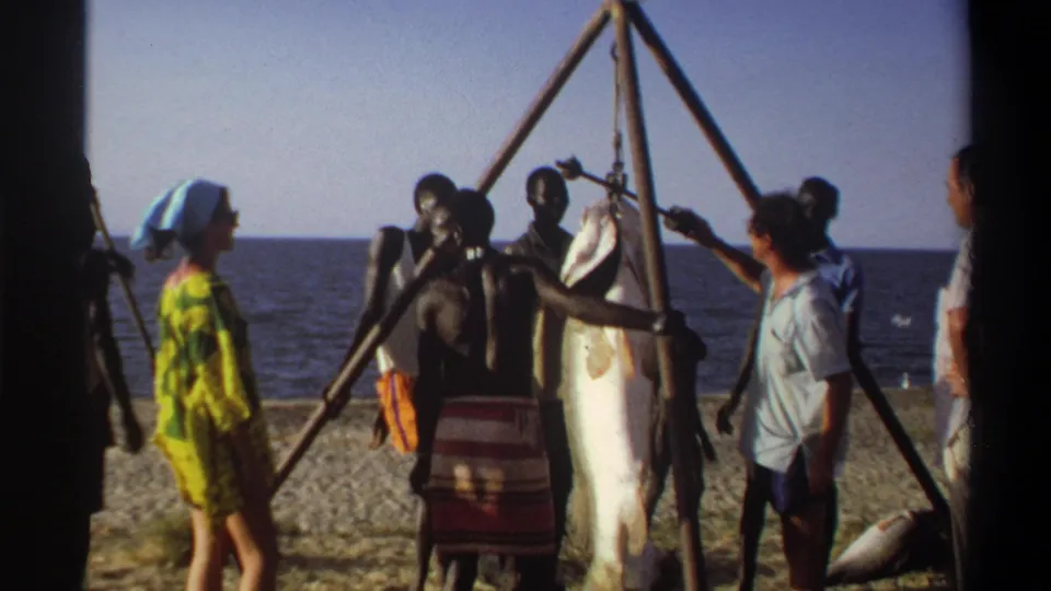 1969 Nigeria beach scene with people gathered around a large fish suspended from a triangular wooden pole rig near the sea.