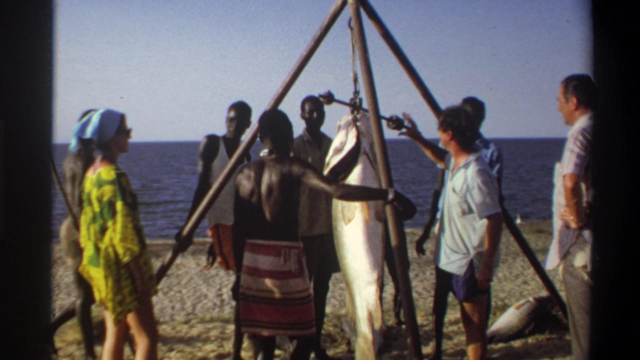 People on a Nigerian beach look toward a large suspended fish on a triangular rig, with the ocean visible behind them.