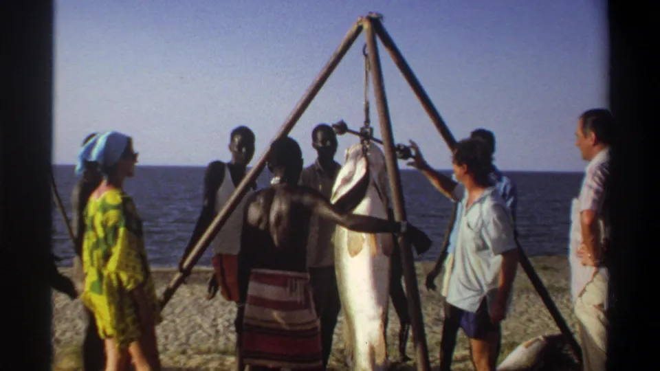 Late-1960s Nigerian beach footage shows people surrounding a large fish hanging from a triangular wooden frame near the sea.