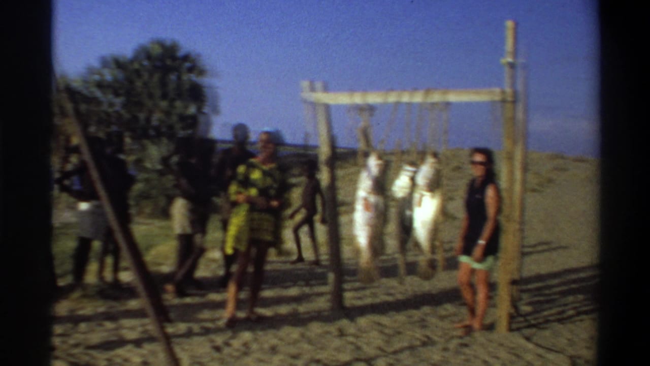 1969 Nigeria 8mm frame showing several people around a beach fish display, with palms and water visible behind.