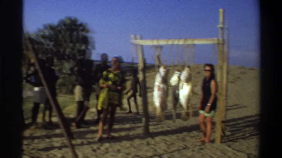 1969 Nigeria 8mm frame showing several people around a beach fish display, with palms and water visible behind.