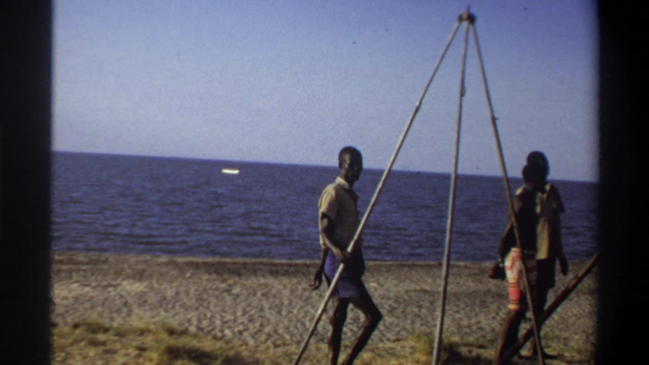 In 1969 Nigeria home-movie footage, two men move along a sandy beach with a tall tripod-like frame and open sea behind them.