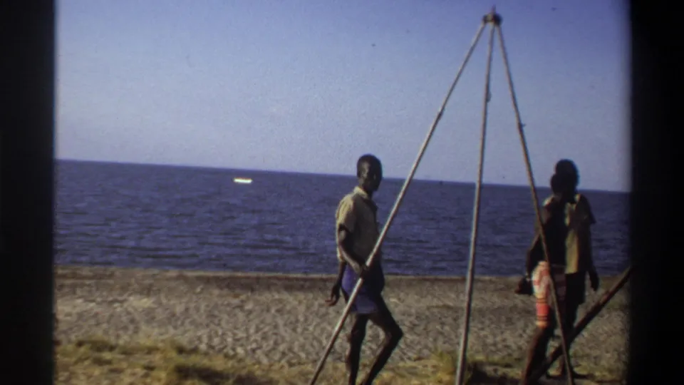 In 1969 Nigeria home-movie footage, two men move along a sandy beach with a tall tripod-like frame and open sea behind them.