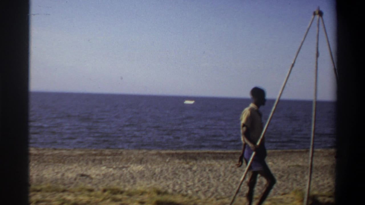 A man walks on a beach beside a tall tripod-like frame with the sea behind him.