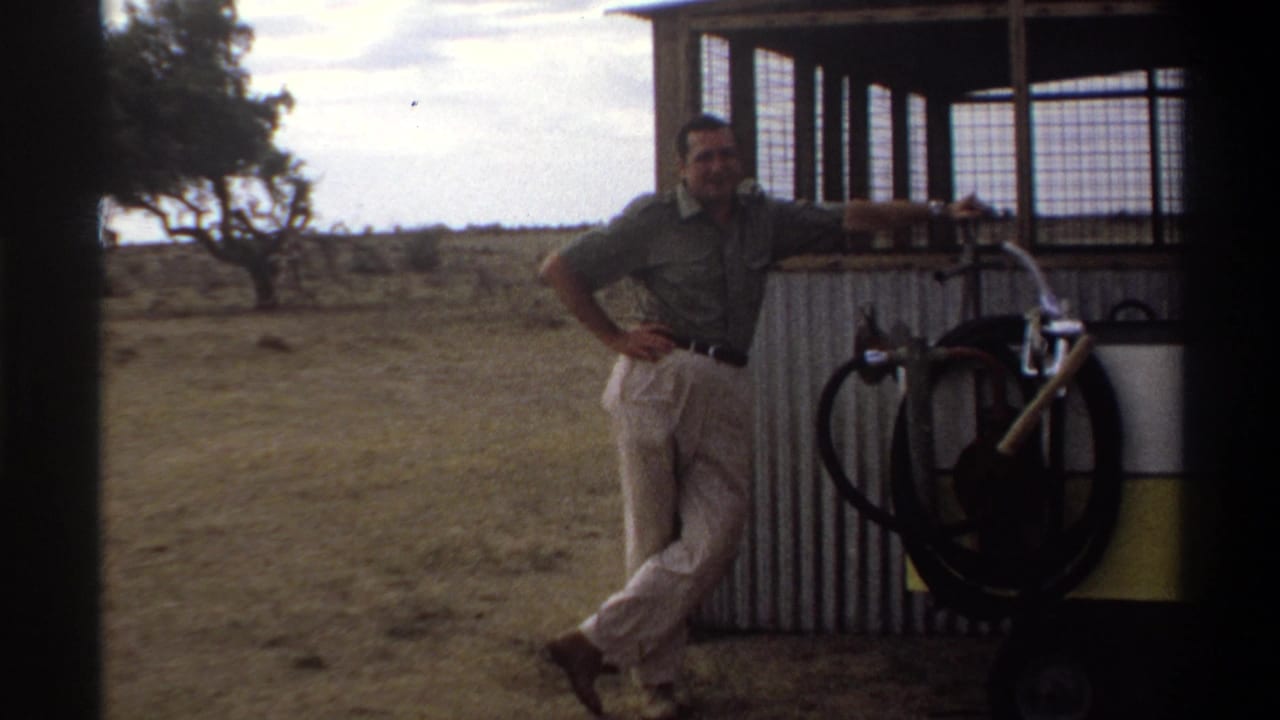 In a 1969 Nigeria outdoor scene, a man in light trousers stands by a corrugated metal structure with a bicycle wheel and open land nearby.