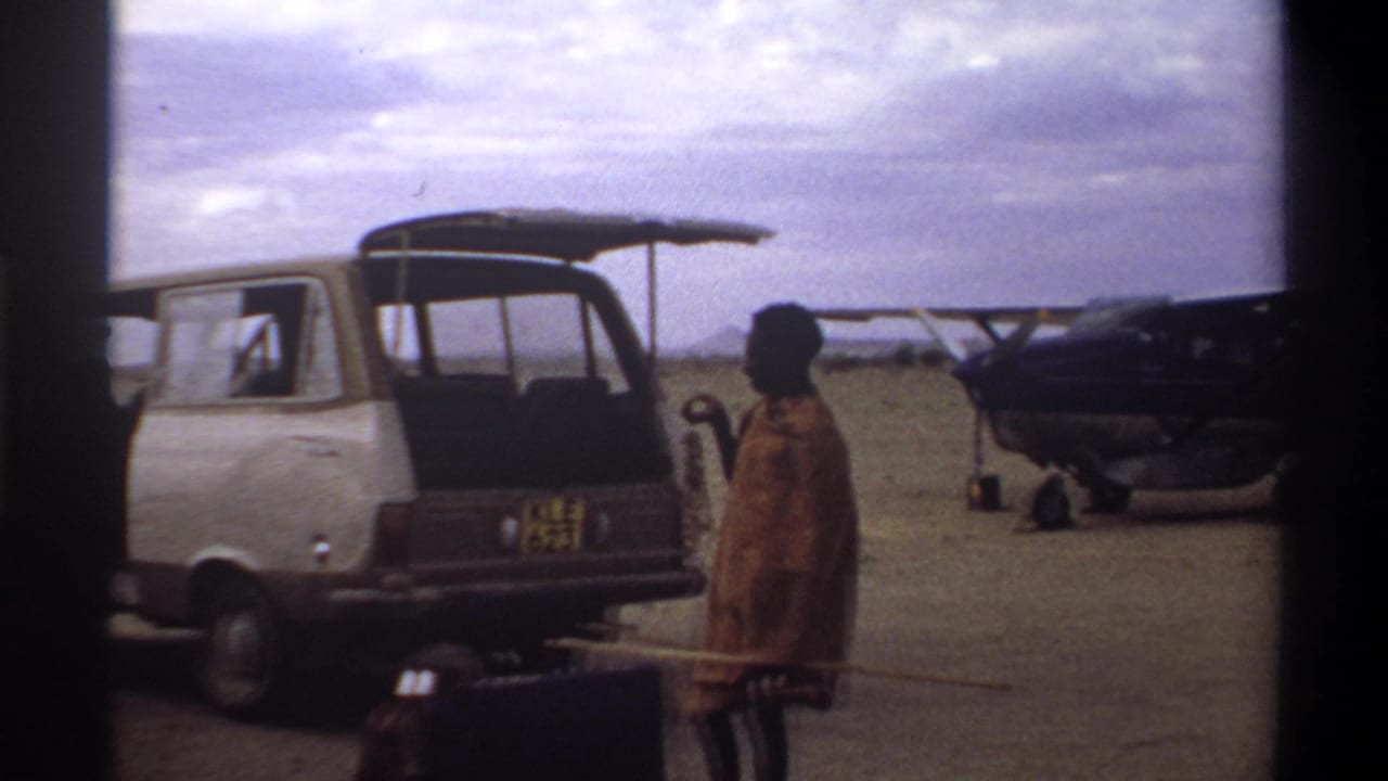 1969 Nigeria home-movie frame showing a van at a dusty airstrip, a person holding a long stick, luggage, and a small propeller plane.