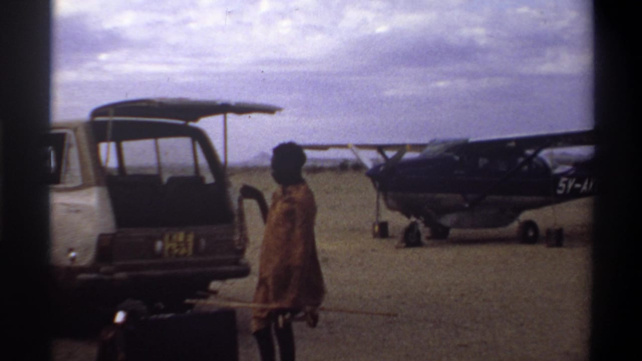 Open van and person in patterned clothing near luggage, with a dark small plane behind them.