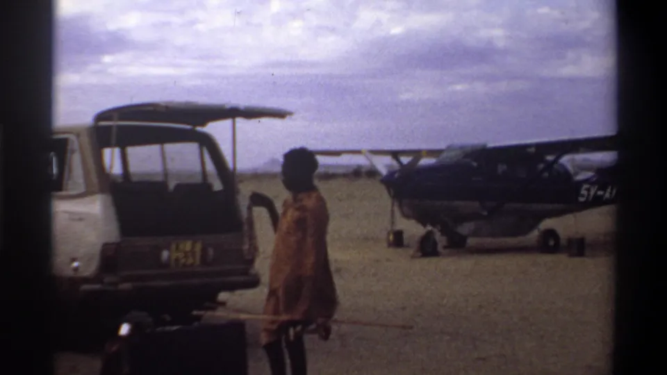 1969 Nigeria travel frame with an open van, a person in patterned clothing, luggage, and a small propeller aircraft on open ground.