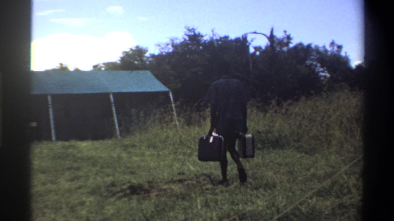 A man seen from behind carries two suitcases across grass toward a green-roofed tent in 1969 Lagos, Nigeria home-movie footage.