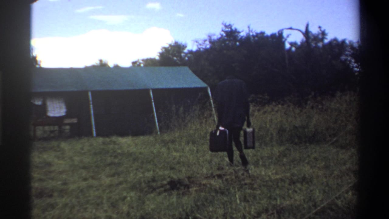 A man in a dark suit carries two suitcases past tall grass and a green-roofed tent in the Lagos, Nigeria 1969 reel.