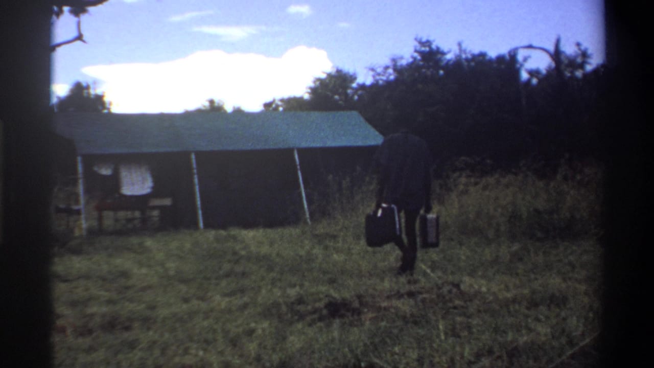 1969 Lagos home-movie frame showing a man in dark clothing carrying two suitcases toward a canvas tent in a grassy field.