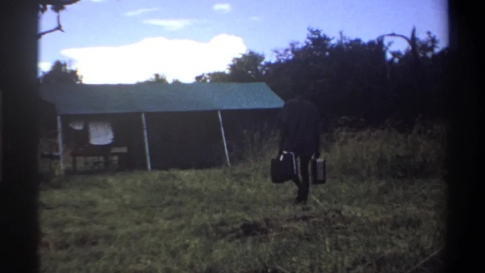 1969 Lagos home-movie frame showing a man in dark clothing carrying two suitcases toward a canvas tent in a grassy field.
