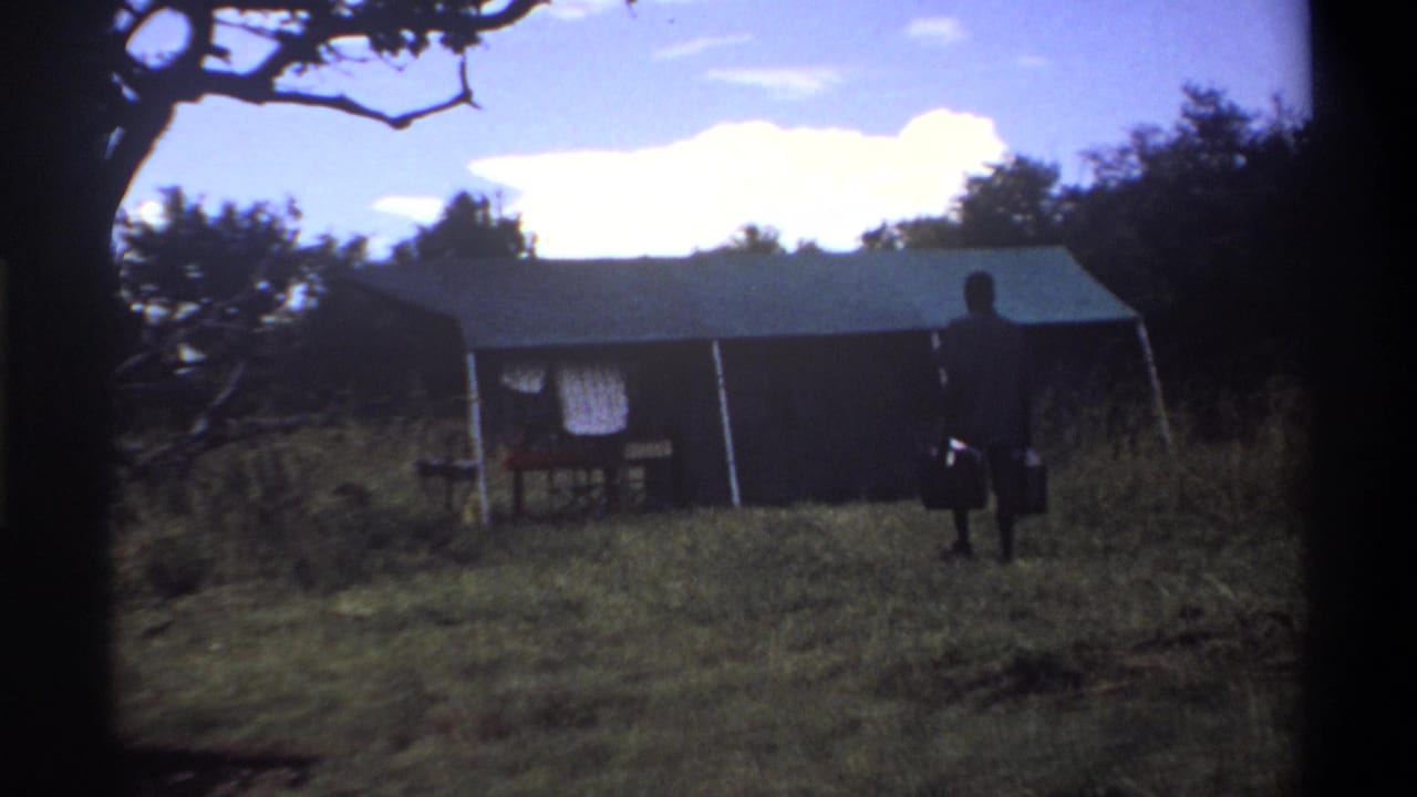 The man appears near the tent in a 1969 Lagos home-movie view, with grass, trees, and a bright sky surrounding the scene.