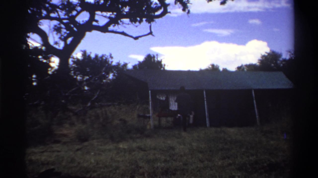 1969 Lagos home-movie frame showing a man near a shaded tent-like shelter beneath a large tree, with bright sky behind.