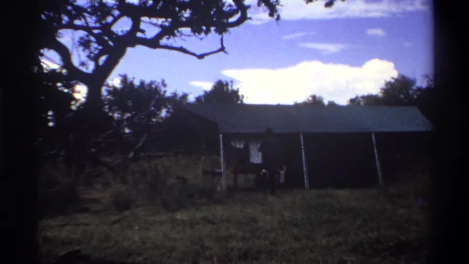 1969 Lagos home-movie frame showing a man near a shaded tent-like shelter beneath a large tree, with bright sky behind.