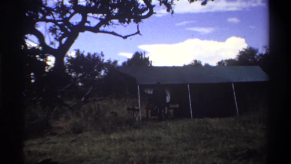 1969 Lagos frame with a dark tent-like structure, tree branches overhead, and a small person partly silhouetted near the opening.