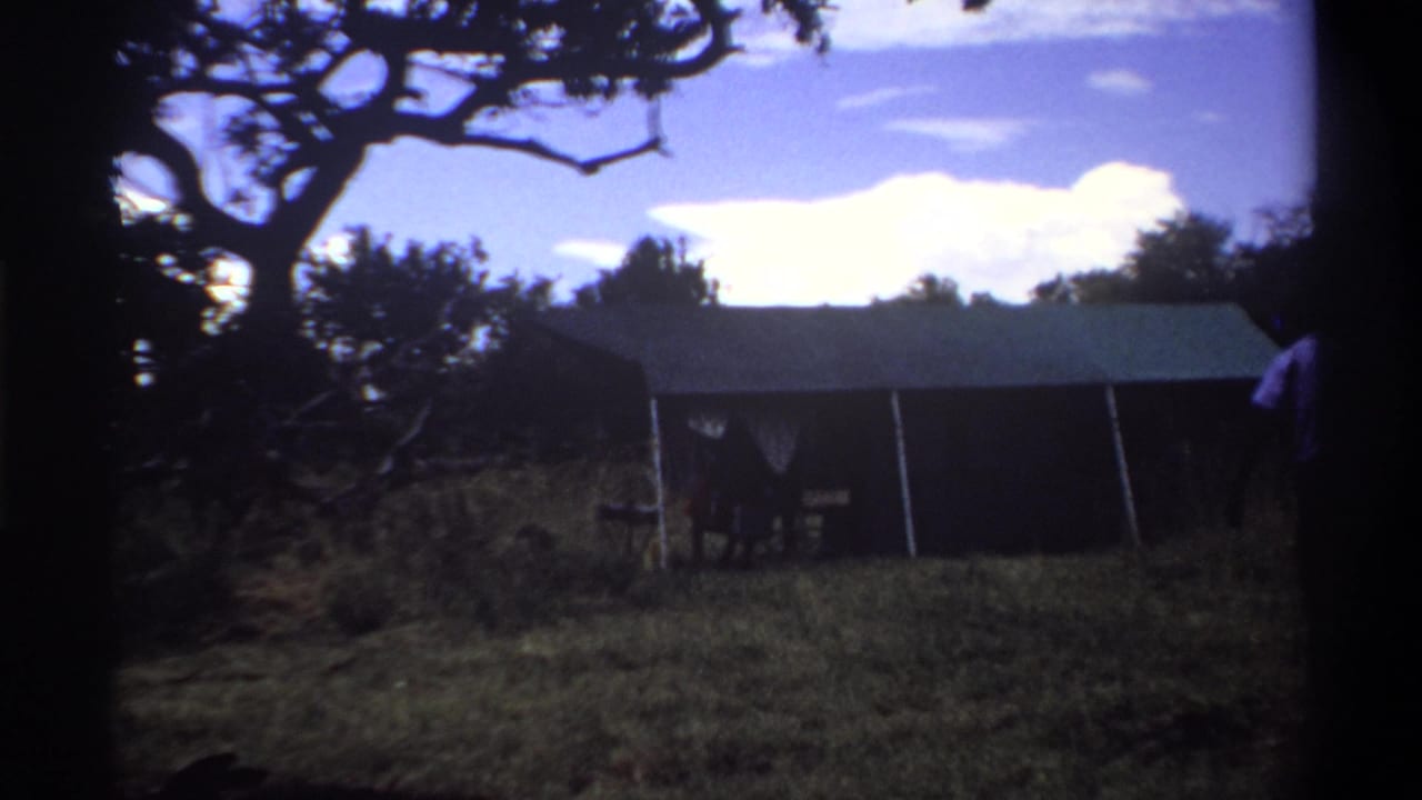 1969 Lagos home-movie frame showing a canvas tent in grass beneath a bright clouded sky, with tree branches silhouetted above.