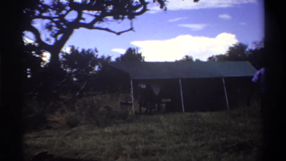 1969 Lagos home-movie frame showing a canvas tent in grass beneath a bright clouded sky, with tree branches silhouetted above.