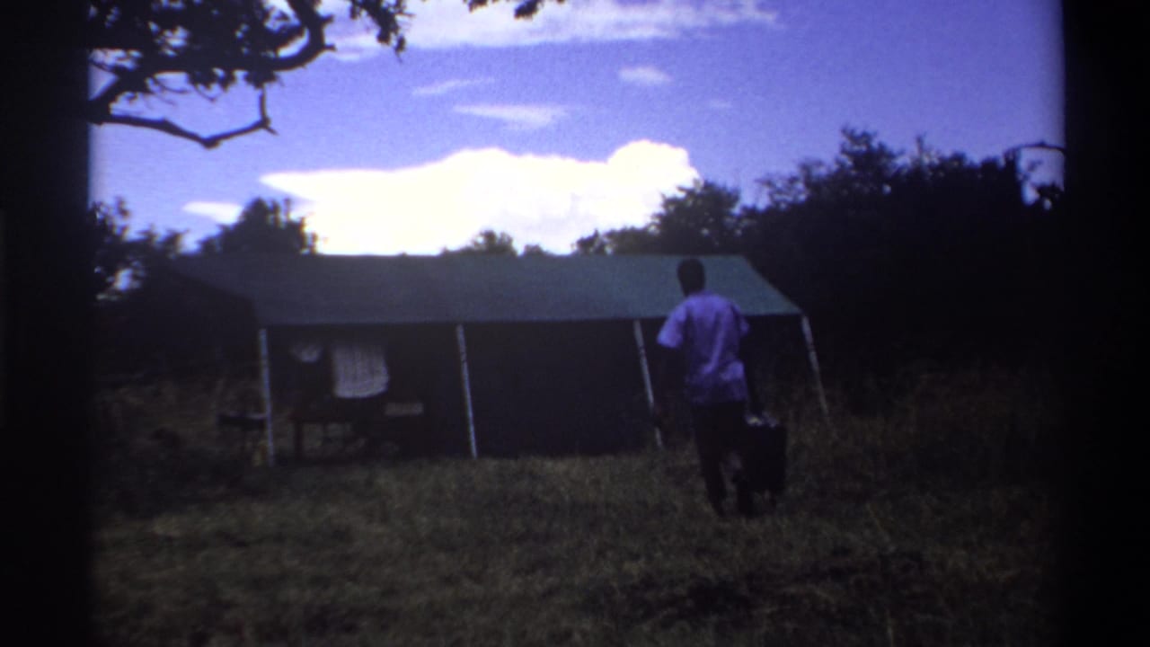 1969 Lagos home-movie frame with a man in a purple shirt carrying dark luggage toward a canvas tent across a grassy field.