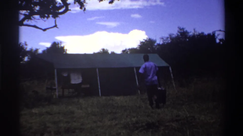 1969 Lagos home-movie frame with a man in a purple shirt carrying dark luggage toward a canvas tent across a grassy field.