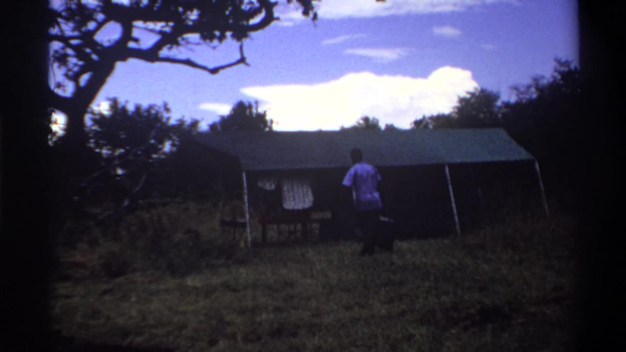 1969 Lagos home-movie frame with a man carrying luggage beside a canvas tent, seen from a shaded grassy clearing.