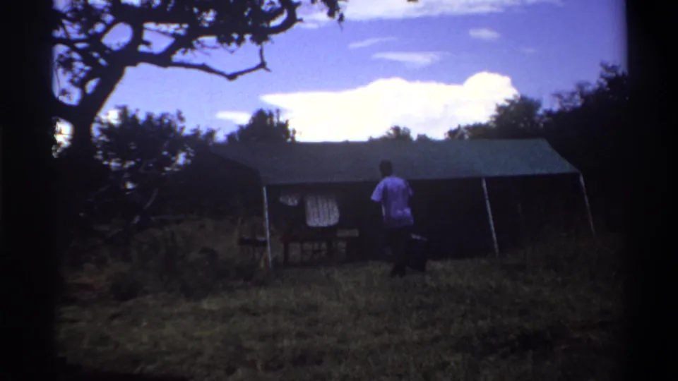 1969 Lagos home-movie frame with a man carrying luggage beside a canvas tent, seen from a shaded grassy clearing.
