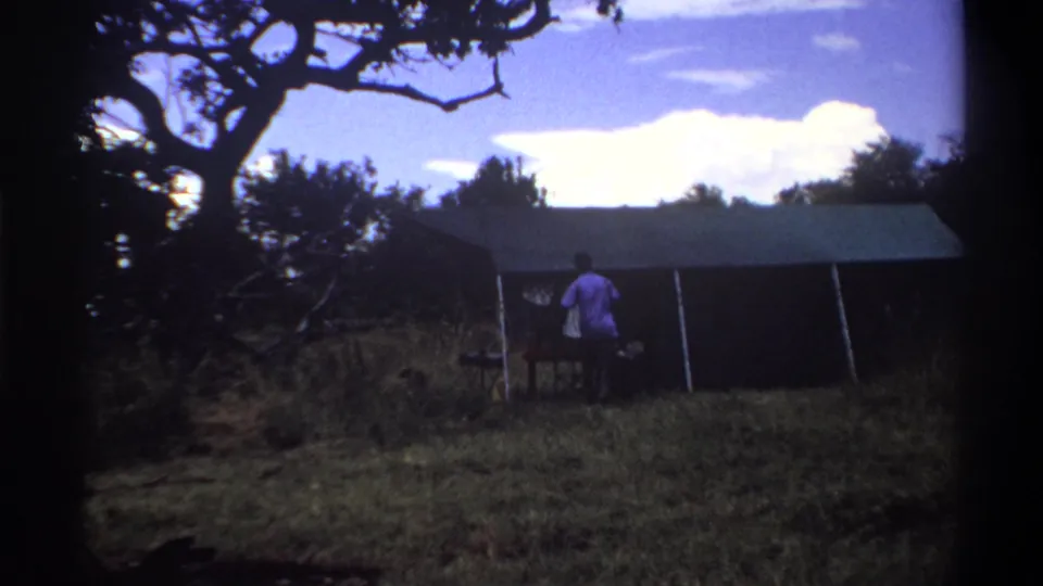 In the 1969 Lagos home-movie frame, the man is closer to the canvas tent entrance with a small table visible beside him.