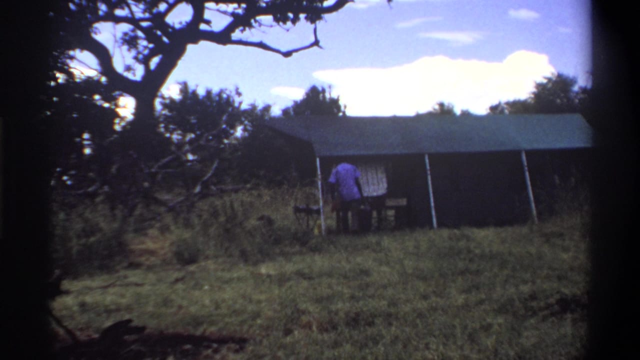 In Lagos, Nigeria, 1969, the man appears to bend near a small table outside the canvas tent in the grassy clearing.