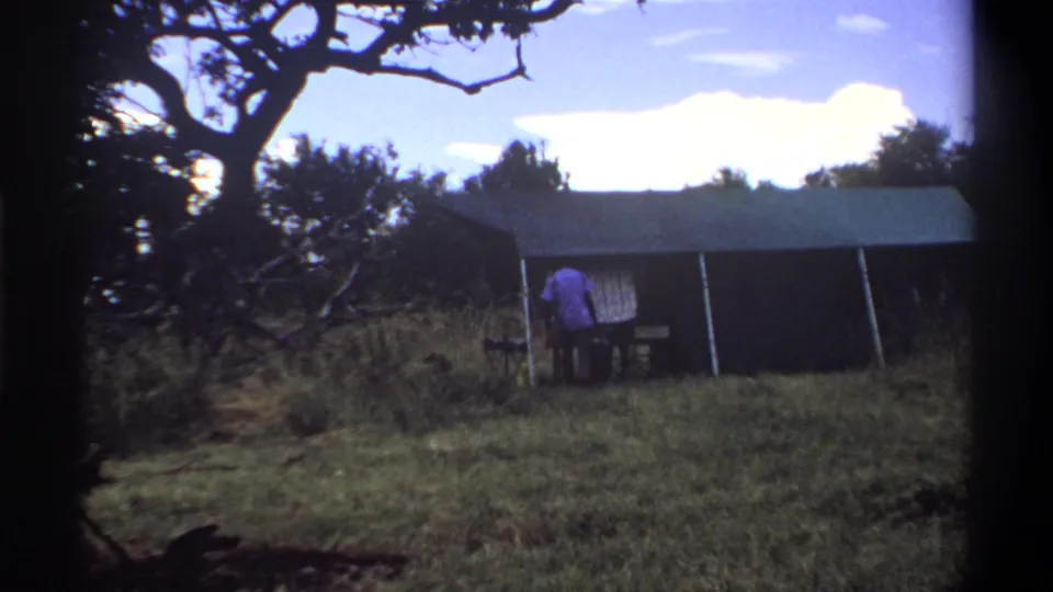 In Lagos, Nigeria, 1969, the man appears to bend near a small table outside the canvas tent in the grassy clearing.