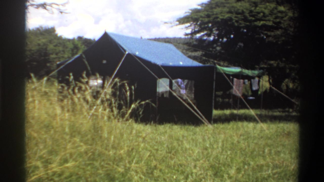 1969 Lagos home-movie frame showing a blue-roofed dark canvas tent, guy ropes, tall grass, trees, and laundry at the side.