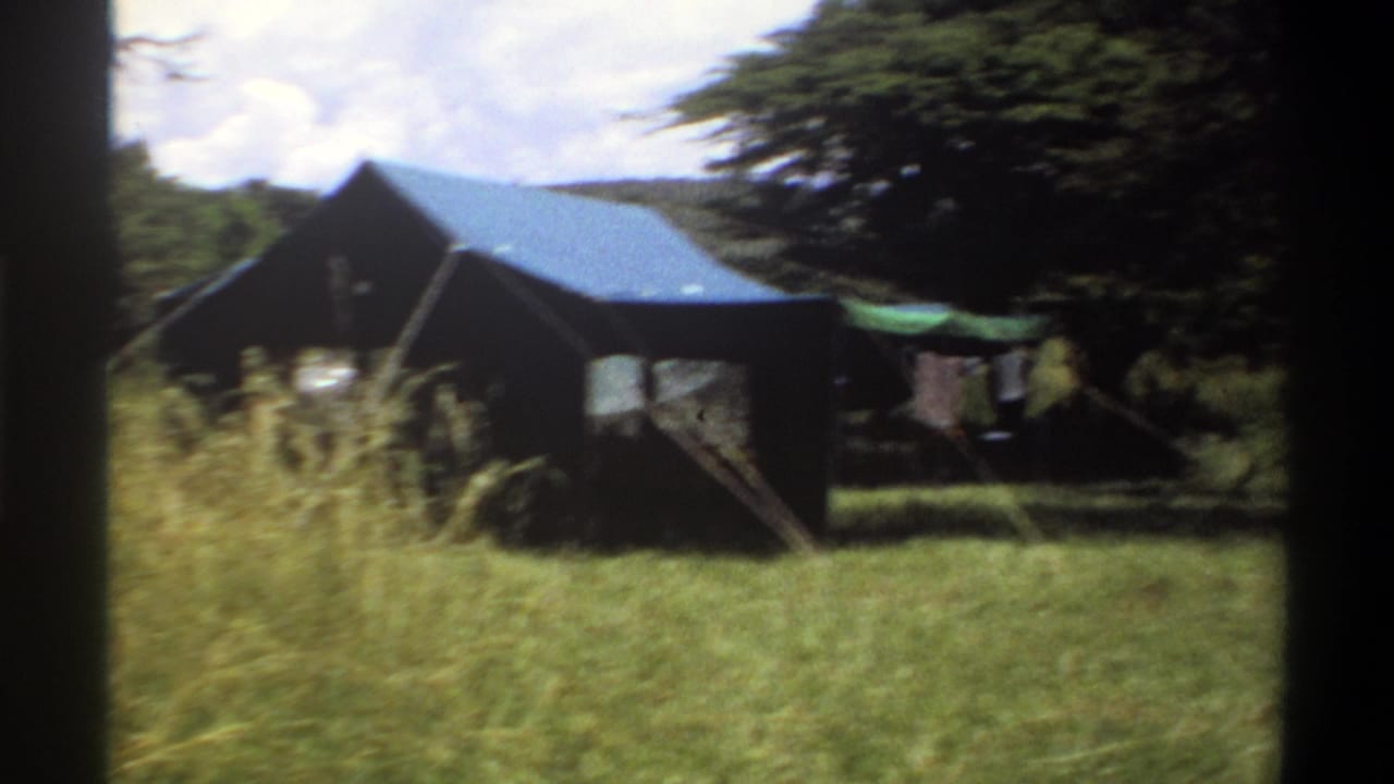 Soft 1969 Lagos 8mm frame showing the blue-roofed tent partly obscured by tall grass, with trees and laundry at the edge.
