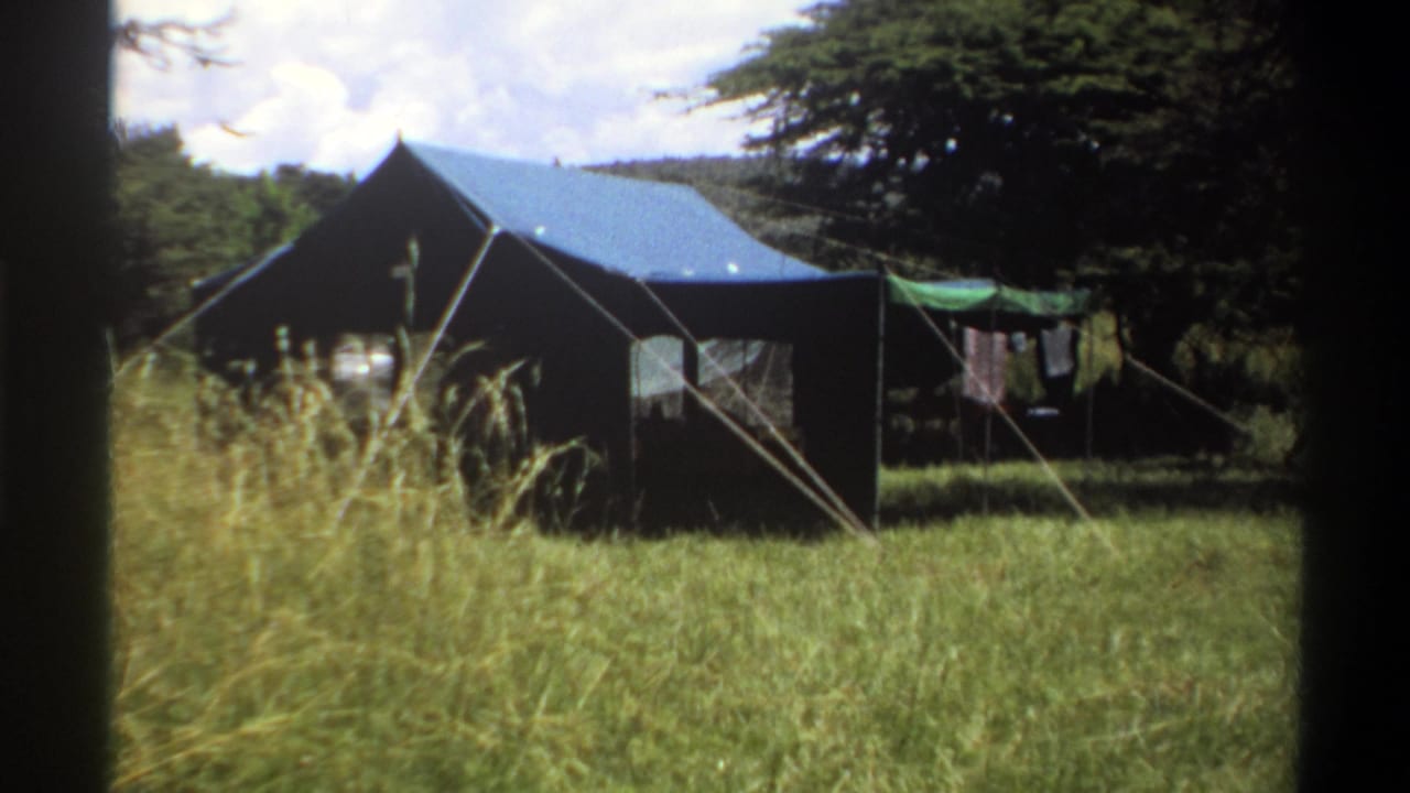1969 Lagos home-movie frame showing the canvas tent from a slightly closer angle, with guy ropes, tall grass, trees, and laundry nearby.