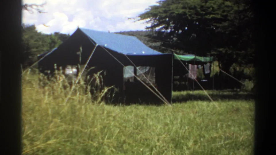 1969 Lagos home-movie frame showing the canvas tent from a slightly closer angle, with guy ropes, tall grass, trees, and laundry nearby.