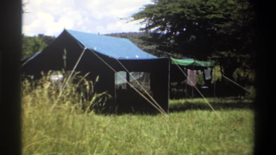 1969 Lagos home-movie frame showing a canvas tent in a grassy clearing with trees, guy ropes, and clothes hanging nearby.