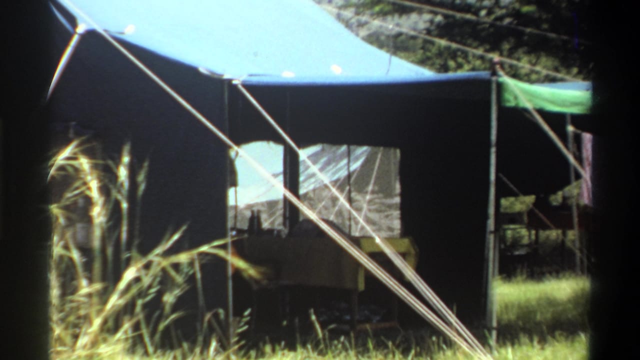 Closer 8mm frame of the Lagos campsite tent, with its window, entrance area, guy ropes, tall grass, and interior table visible.