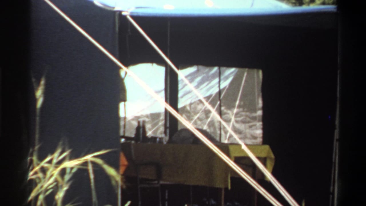 Final close frame of the 1969 Lagos tent, showing the window, dark canvas wall, guy ropes, tall grass, and furniture inside the opening.
