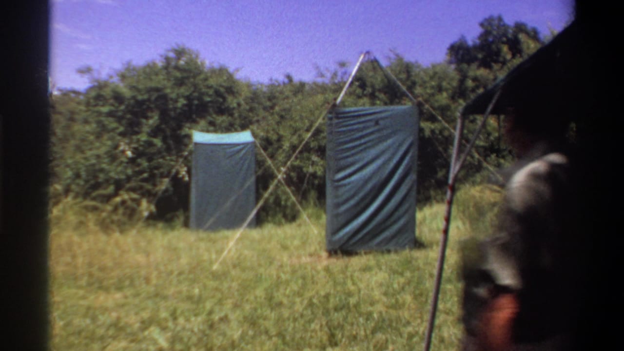Man partly entering a grassy field view toward two tarp shelters under a blue sky.