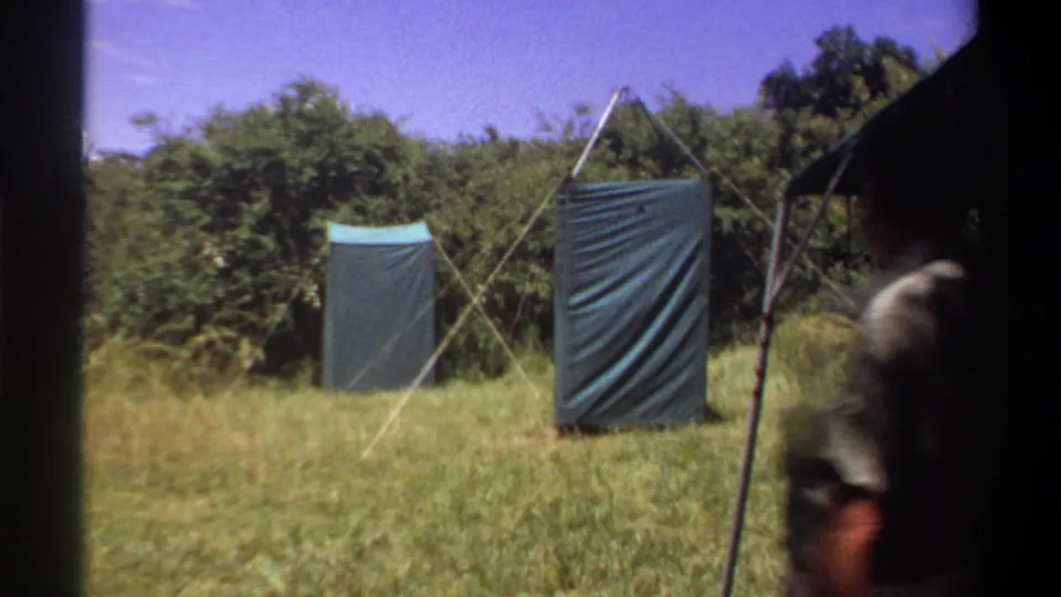 1969 Lagos home-movie frame showing a man partly obscured at the edge of a grassy field with two blue-green tarp shelters ahead.